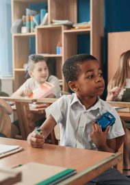 Curious boy on the foreground holding a toy car and a battery. Children sitting at their desks at the lesson
