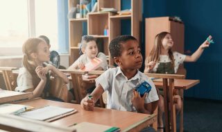 Curious boy on the foreground holding a toy car and a battery. Children sitting at their desks at the lesson
